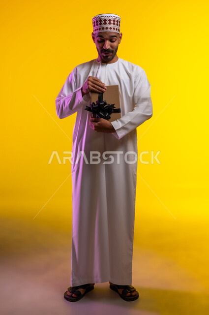 ايماءات وجه تدل على الحماس لمعرفه ما بداخل صندوق الهدايا، بورتريه لرجل عربي خليجي عماني يرتدي الدشداشة والكمه يمسك بيده هديه كرتونيه يقوم بمحاوله فتحها، تعابير فرح وسرور واكتشاف المفاجات السعيده، هدايا عيد الاب ومناسبات سعيده، خلفيه برتقاليه