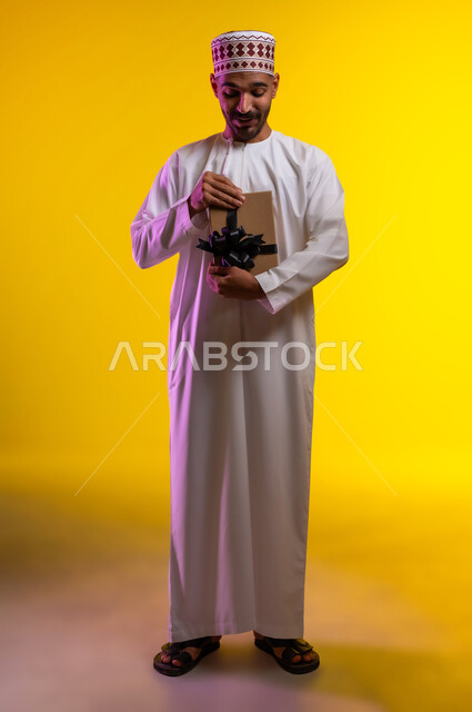 ايماءات وجه تدل على الحماس لمعرفه ما بداخل صندوق الهدايا، بورتريه لرجل عربي خليجي عماني يرتدي الدشداشة والكمه يمسك بيده هديه كرتونيه يقوم بمحاوله فتحها، تعابير فرح وسرور واكتشاف المفاجات السعيده، هدايا عيد الاب ومناسبات سعيده، خلفيه برتقاليه
