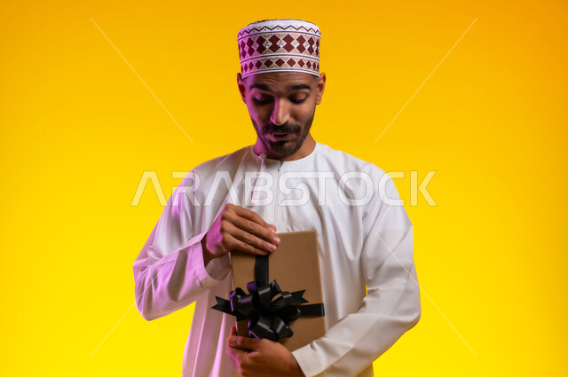 ايماءات وجه تدل على الحماس لمعرفه ما بداخل صندوق الهدايا، بورتريه لرجل عربي خليجي عماني يرتدي الدشداشة والكمه يمسك بيده هديه كرتونيه يقوم بمحاوله فتحها، تعابير فرح وسرور واكتشاف المفاجات السعيده، هدايا عيد الاب ومناسبات سعيده، خلفيه برتقاليه