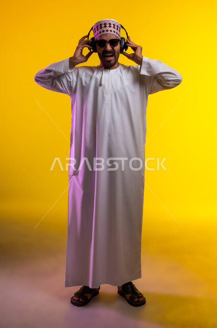 مواهب غنائية فنية، إيماءات وتعابير الاندماج مع الأغنية، بورتريه لرجل عربي خليجي عماني يرتدي الدشداشه والكمه يستمع الى الموسيقى من خلال سماعه الرأس، ايماءات تدل على الانصات والإستماع للأغاني بإستخدام الهيدفون، التفاعل مع الانغام الموسيقية، خلفية صفراء