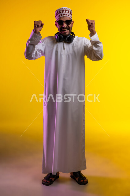 ايماءات وجه تدل على الحماس الزائد، بورتريه لرجل عربي خليجي عماني يرتدي الدشداشة والكمه يضع سماعه هيدفون حول رقبته و يرفع يده للأعلى للتشجيع، تعابير فرح وسروروالشعور بالسعادة والحماس للفوز وتحقيق هدف ما، خلفية برتقاليه