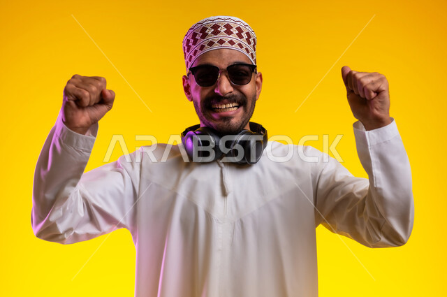 ايماءات وجه تدل على الحماس الزائد، بورتريه لرجل عربي خليجي عماني يرتدي الدشداشة والكمه يضع سماعه هيدفون حول رقبته و يرفع يده للأعلى للتشجيع، تعابير فرح وسروروالشعور بالسعادة والحماس للفوز وتحقيق هدف ما، خلفية برتقاليه