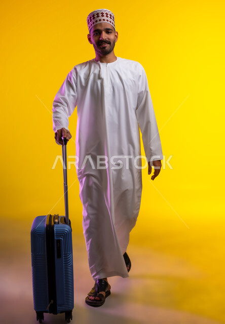 الاستعداد لوقت الرحلة، مفهوم السياحة والسفر، قضاء أوقات ممتعة في الإجازة, بورتريه لشاب عربي خليجي عماني يرتدي الدشداشة و الكمه يمسك حقيبة السفر بيده, النظر الى شيء ما بتعابير الحماس والسرور, خلفيه برتقاليه