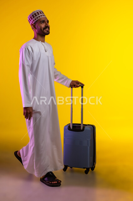 الاستعداد لوقت الرحلة، مفهوم السياحة والسفر، قضاء أوقات ممتعة في الإجازة, بورتريه لشاب عربي خليجي عماني يرتدي الدشداشة و الكمه يمسك حقيبة السفر بيده, النظر الى شيء ما بتعابير الحماس والسرور, خلفيه برتقاليه