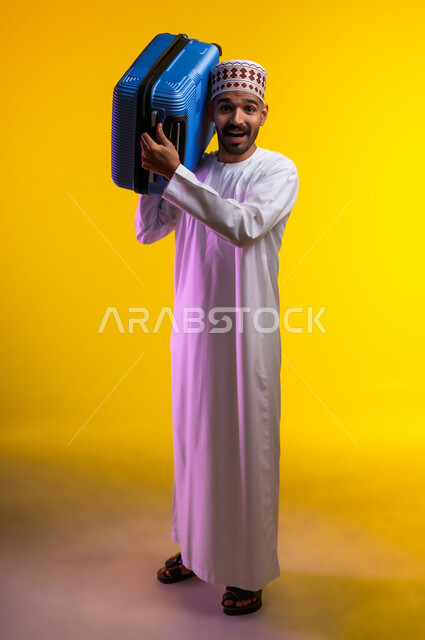 الاستعداد لوقت الرحلة، مفهوم السياحة والسفر، قضاء أوقات ممتعة في الإجازة, بورتريه لشاب عربي خليجي عماني يرتدي الدشداشة و الكمه يمسك حقيبة السفر بيده, النظر الى شيء ما بتعابير الحماس والسرور, خلفيه برتقاليه