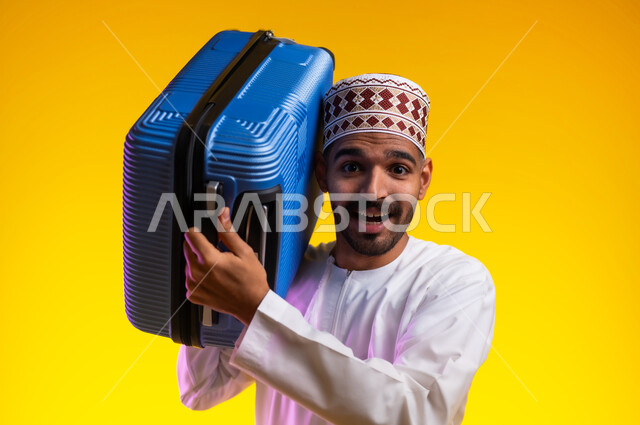الاستعداد لوقت الرحلة، مفهوم السياحة والسفر، قضاء أوقات ممتعة في الإجازة, بورتريه لشاب عربي خليجي عماني يرتدي الدشداشة و الكمه يمسك حقيبة السفر بيده, النظر الى شيء ما بتعابير الحماس والسرور, خلفيه برتقاليه