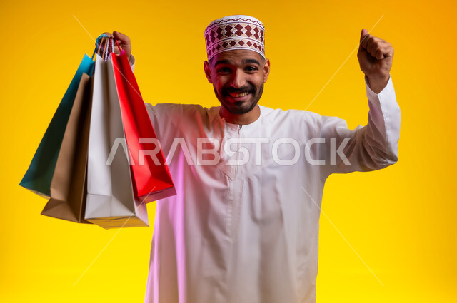 اقتناء المستلزمات والهدايا، الاستفادة من عروض وتخفيضات الجمعة البيضاء، بورتريه لرجل عربي خليجي عماني مبتسم يرتدي الدشداشه والكمه يحمل بيده أكياس تسوق ورقية ملونة ينظر ويشير بيديه إلى الكاميرا بإيماءات السرور والسعادة والحماس، خلفية برتقاليه