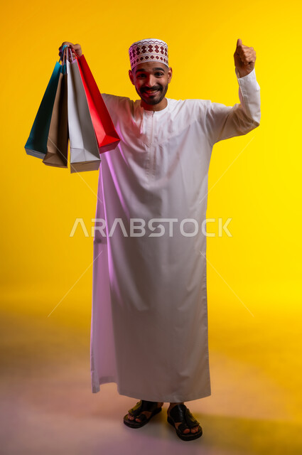 اقتناء المستلزمات والهدايا، الاستفادة من عروض وتخفيضات الجمعة البيضاء، بورتريه لرجل عربي خليجي عماني مبتسم يرتدي الدشداشه والكمه يحمل بيده أكياس تسوق ورقية ملونة ينظر ويشير بيديه إلى الكاميرا بإيماءات السرور والسعادة والحماس، خلفية برتقاليه