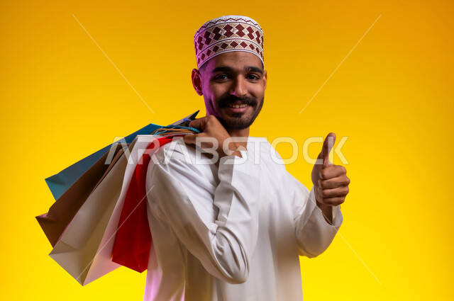 ايماءات تدل على الموافقة والقبول، بورتريه لرجل عربي خليجي عماني يرتدي الدشداشة والكمه يشير باصبع الابهام للاعلى تظهر عليه تعابير الفرح والسرور يحمل بيده اكياس التسوق الملونه، الشراء من الاسواق والمحال التجاريه، خلفية برتقاليه