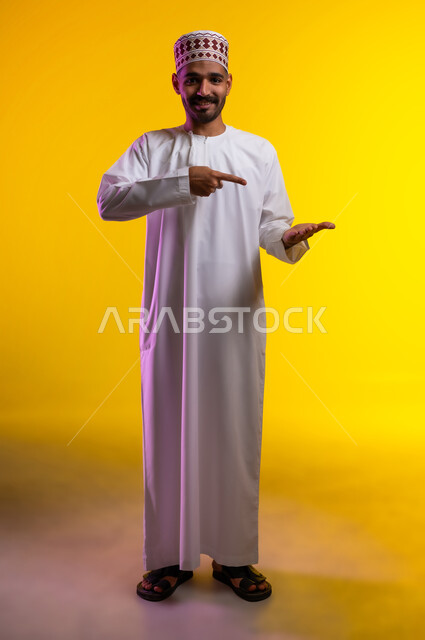 تعابير الفرح والسرور والاشاره الى شئ ما، بورتريه لشاب عربي خليجي عماني مبتسم يرتدي الدشداشة و الكمه يشير بإصبع السبابة ليده الأخرى، الامساك بعنصر ما بإيماءات الاعجاب، الانتباه والتركيز، خلفية برتقاليه