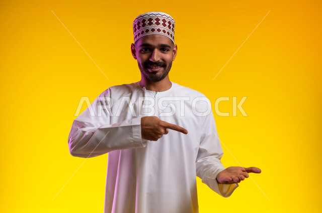 تعابير الفرح والسرور والاشاره الى شئ ما، بورتريه لشاب عربي خليجي عماني مبتسم يرتدي الدشداشة و الكمه يشير بإصبع السبابة ليده الأخرى، الامساك بعنصر ما بإيماءات الاعجاب، الانتباه والتركيز، خلفية برتقاليه