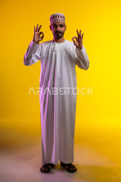 التعبير عن الجودة والإتقان, بورتريه لرجل عربي خليجي عماني يرتدي الدشداشة والكمه  يرفع يده للأعلى  بإيماءات الرضا والقبول والأعجاب, النظر الى الكاميرا بتعابير السرور, صورة الجسم بالطول الكامل, خلفية برتقاليه