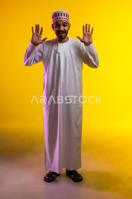 Expressions of rejection and abstention, a portrait of a young Arab Gulf Omani man wearing a dishdasha and a cuff, pointing forward with his hands with gestures of prohibition and stopping doing something, feeling astonished and shocked, orange background.