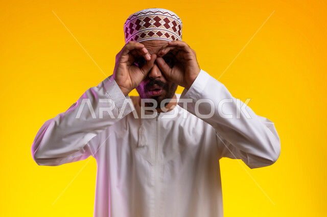مفهوم التخطيط و الرؤية المستقبلية، بورتريه لرجل عربي خليجي عماني يرتدي الدشداشة والكمه يضع يده على عينيه بشكل عدسة بإيماءات البحث والاستكشاف، النظر الى مكان بعيد وتقريب الاشياء البعيده، خلفيه برتقاليه