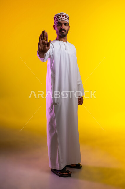 The command to stop doing something, expressions of rejection and abstinence, a portrait of a young Arab Gulf Omani man wearing a dishdasha and a cuff, raising his hand with prohibitive gestures, feeling annoyed and angry, orange background.
