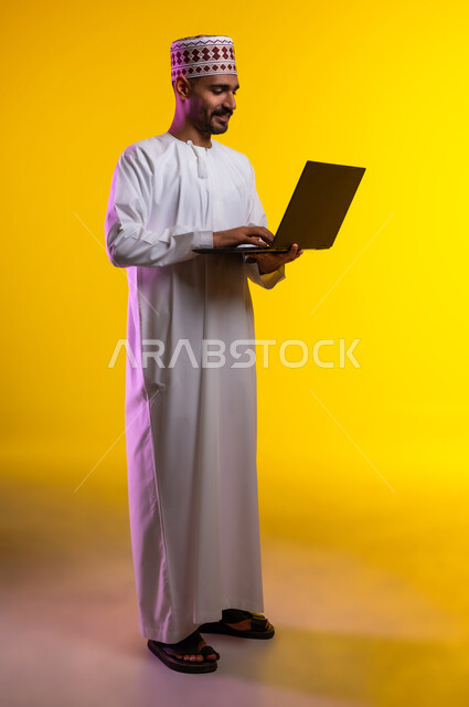 مفهوم العمل والاجتهاد وانجاز الاعمال، بورتريه لشاب عربي خليجي عماني يرتدي الدشداشة والكمه يقف ويمسك بيده جهاز الابتوب المحمول، تبادل الثقافات والاراء، استخدام التقنيات الحديثه والعمل عن بعد، خلفية صفراء