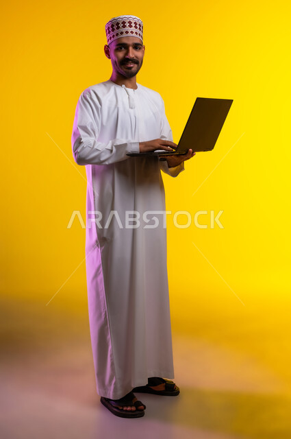 مفهوم العمل والاجتهاد وانجاز الاعمال، بورتريه لشاب عربي خليجي عماني يرتدي الدشداشة والكمه يقف ويمسك بيده جهاز الابتوب المحمول، تبادل الثقافات والاراء، استخدام التقنيات الحديثه والعمل عن بعد، خلفية صفراء