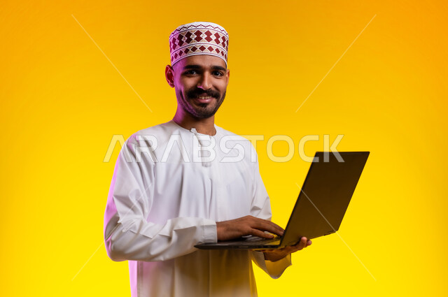 مفهوم العمل والاجتهاد وانجاز الاعمال، بورتريه لشاب عربي خليجي عماني يرتدي الدشداشة والكمه يقف ويمسك بيده جهاز الابتوب المحمول، تبادل الثقافات والاراء، استخدام التقنيات الحديثه والعمل عن بعد، خلفية صفراء