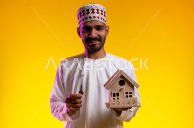 اسكان وتمليك، ايماءات تعبر عن السعادة بشراء عقار جديد, بورتريه لرجل عربي خليجي عماني يرتدي الدشداشه والكمه يحمل بيده مجسما لمنزل صغير ومفتاح البيت الجديد، امتلاك سكن حديث والاستقرار العائلي، خلفية برتقاليه