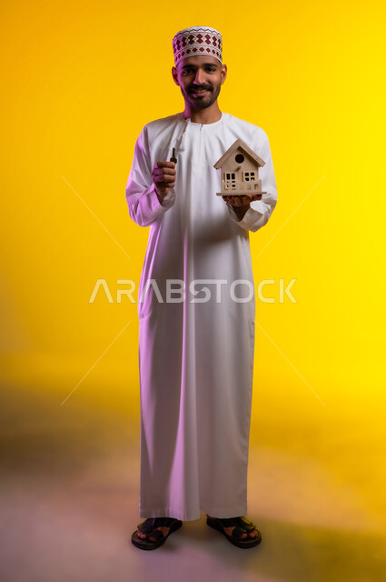 اسكان وتمليك، ايماءات تعبر عن السعادة بشراء عقار جديد, بورتريه لرجل عربي خليجي عماني يرتدي الدشداشه والكمه يحمل بيده مجسما لمنزل صغير ومفتاح البيت الجديد، امتلاك سكن حديث والاستقرار العائلي، خلفية برتقاليه