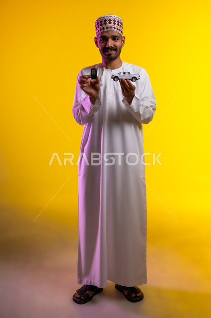 السعاده بامتلاك سياره حديثه، الفضول بتجربه قياده المركبات الجديده، بورتريه لرجل عربي خليجي عماني يرتدي الدشداشه والكمه يحمل بيده مجسما لسياره مصغره وجهاز الريموت الخاص بالتحكم عن بعد  بايماءات الفرح والسرور، موديلات وطرازات، خلفيه برتقاليه
