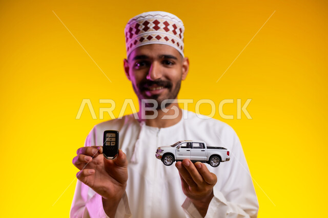 Happiness in owning a modern car, curiosity in the experience of driving new vehicles, a portrait of an Arab Gulf Omani man wearing a dishdasha and a cuff holding in his hand a model of a miniature car and the remote control device with gestures of joy and pleasure, models and styles, orange background