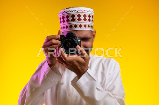 فوتوغرافر محترف يستعد لالتقاط الصور، النظر من خلال عدسات الكاميرا بايماءات الثقه بالنفس والجديه، التقاط صور بدقه عاليه، بورتريه لرجل عربي خليجي عماني يرتدي الدشداشه والكمه يقوم بالتقاط الصور الفوتوغرافيه بتركيز وتمعن، صور فوتوغرافيه حديثه، خلفيه صفراء