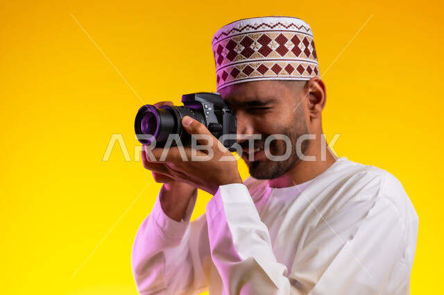 فوتوغرافر محترف يستعد لالتقاط الصور، النظر من خلال عدسات الكاميرا بايماءات الثقه بالنفس والجديه، التقاط صور بدقه عاليه، بورتريه لرجل عربي خليجي عماني يرتدي الدشداشه والكمه يقوم بالتقاط الصور الفوتوغرافيه بتركيز وتمعن، صور فوتوغرافيه حديثه، خلفيه صفراء
