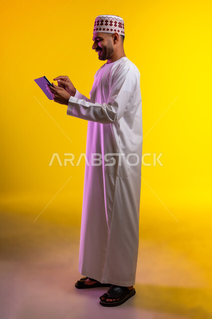 مفهوم العمل والاجتهاد وانجاز الاعمال، بورتريه لشاب عربي خليجي عماني يرتدي الدشداشة والكمه يقف ويمسك بيده جهاز التابلت المحمول، تبادل الثقافات والاراء واستخدام التقنيات الحديثه والعمل عن بعد، خلفية صفراء
