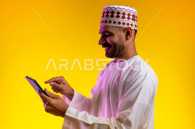 مفهوم العمل والاجتهاد وانجاز الاعمال، بورتريه لشاب عربي خليجي عماني يرتدي الدشداشة والكمه يقف ويمسك بيده جهاز التابلت المحمول، تبادل الثقافات والاراء واستخدام التقنيات الحديثه والعمل عن بعد، خلفية صفراء