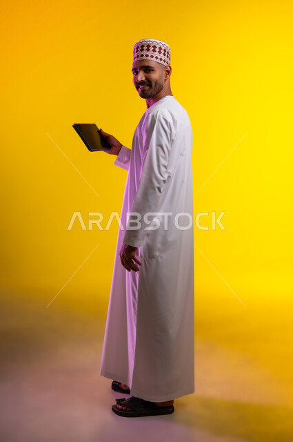 مفهوم العمل والاجتهاد وانجاز الاعمال، بورتريه لشاب عربي خليجي عماني يرتدي الدشداشة والكمه يقف ويمسك بيده جهاز التابلت المحمول، تبادل الثقافات والاراء واستخدام التقنيات الحديثه والعمل عن بعد، خلفية صفراء