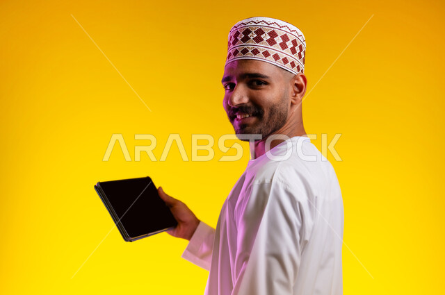 مفهوم العمل والاجتهاد وانجاز الاعمال، بورتريه لشاب عربي خليجي عماني يرتدي الدشداشة والكمه يقف ويمسك بيده جهاز التابلت المحمول، تبادل الثقافات والاراء واستخدام التقنيات الحديثه والعمل عن بعد، خلفية صفراء