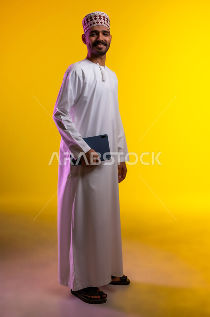 مفهوم العمل والاجتهاد وانجاز الاعمال، بورتريه لشاب عربي خليجي عماني يرتدي الدشداشة والكمه يقف ويمسك بيده جهاز التابلت المحمول، تبادل الثقافات والاراء واستخدام التقنيات الحديثه والعمل عن بعد، خلفية صفراء