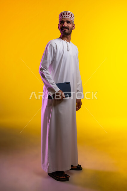 مفهوم العمل والاجتهاد وانجاز الاعمال، بورتريه لشاب عربي خليجي عماني يرتدي الدشداشة والكمه يقف ويمسك بيده جهاز التابلت المحمول، تبادل الثقافات والاراء واستخدام التقنيات الحديثه والعمل عن بعد، خلفية صفراء