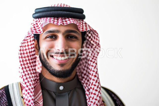 مفهوم الرجولة والأناقه، الاهتمام بالمظهر الخارجي، بورتريه مقرب لرجل عربي خليجي سعودي مبتسم يرتدي الشماغ والثوب التقليدي ينظر إلى الكاميرا بإيماءات السرور والفرح، خلفية بيضاء