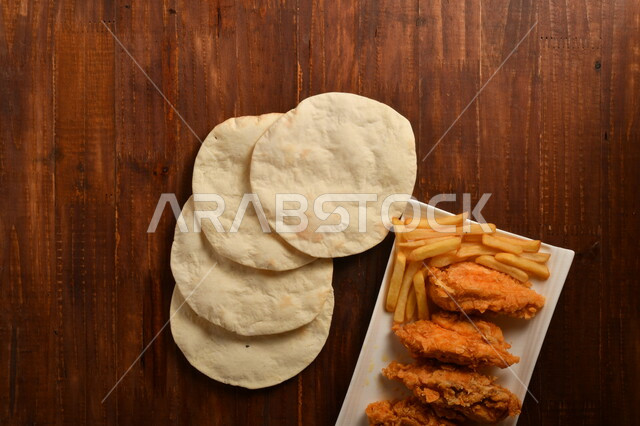 طبق لذيذ مكون من قطع من الدجاج المقلية الكرسبي اللذيذة المغلفة بالبقسماط ، مع بطاطا المقلية و الخبز الابيض العربي ، موضوع على سطح خشبي