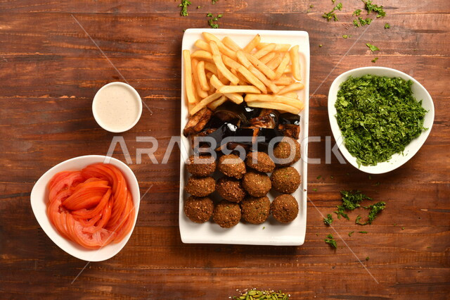 طبق ابيض لذيذ يحتوي على مجموعة من اقراص الفلافل الشهية و البطاطا المقلية ، مع مجموعة من المقبلات بجانبه ، طبق بقدونس ، طبق طماطم ، صوص طحينة ، مأكولات عربي شامية ، موضوع على سطح خشبي