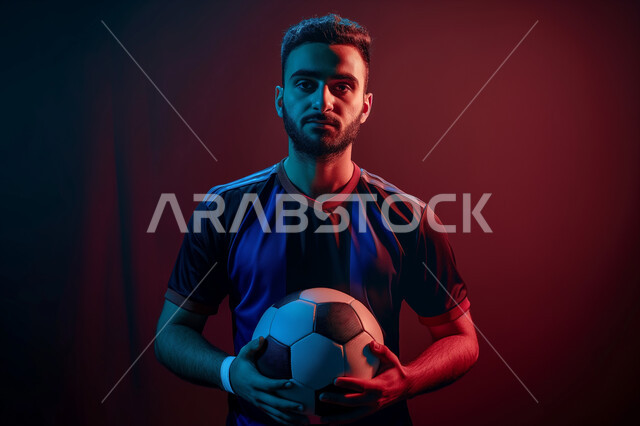 الوقوف باستقامة والنظر الى الكاميرا، اتقان الرياضات السعودية الشبابية، ممارسة الهوايات الرياضية، بورتريه مقرب لشاب عربي خليجي سعودي يرتدي الملابس الرياضية يمسك بيده كرة القدم بتعابير الجدية والثقة بالنفس، خلفية سوداء مع اضاءة حمراء