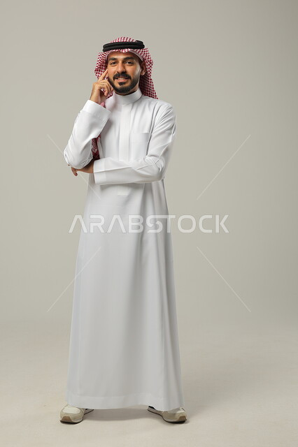 التأمل والتخطيط للمستقبل، التركيز وشرود الذهن والتمعن، إيجاد حل لمشكلة ما، بورتريه لشاب عربي خليجي سعودي مبتسم يرتدي الثوب والشماغ التقليدي يضع يده على ذقنه و ينظر إلى الكاميرا بإيماءات التفكير، صورة الجسم بالطول الكامل، خلفية رمادية