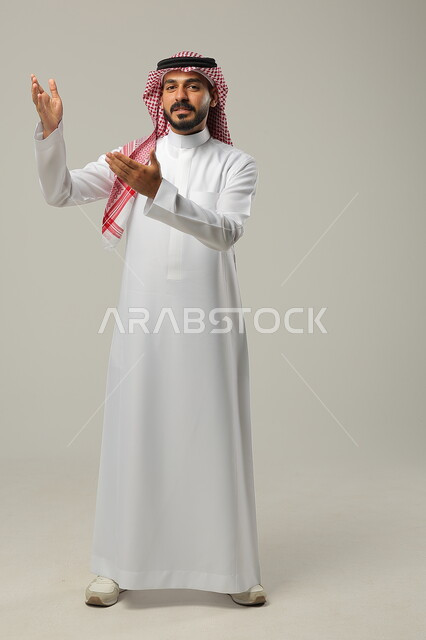 الإشارة الى شخص ما بحركات التواجد هنا, ايماءات التأكيد وتحديد الشيء المقابل, بورتريه لرجل عربي خليجي سعودي يرتدي الثوب التقليدي والشماغ يرفع يديه للأمام بتعابير السرور, صورة الجسم بالطول الكامل, خلفية رمادية