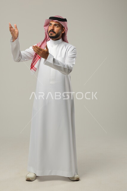 الإشارة الى شخص ما بحركات التواجد هنا, ايماءات التأكيد وتحديد الشيء المقابل, بورتريه لرجل عربي خليجي سعودي يرتدي الثوب التقليدي والشماغ يرفع يديه للأمام بتعابير السرور, صورة الجسم بالطول الكامل, خلفية رمادية