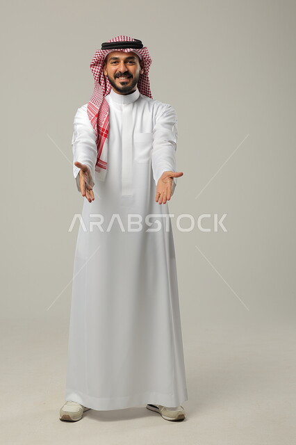 ايماءات التأكيد وتحديد الشيء المقابل, الإشارة الى شخص ما بحركات التواجد هنا, بورتريه لرجل عربي خليجي سعودي يرتدي الثوب التقليدي والشماغ يرفع يديه للأمام بتعابير السرور, صورة الجسم بالطول الكامل, خلفية رمادية