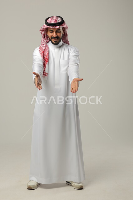 ايماءات التأكيد وتحديد الشيء المقابل, الإشارة الى شخص ما بحركات التواجد هنا, بورتريه لرجل عربي خليجي سعودي يرتدي الثوب التقليدي والشماغ يرفع يديه للأمام بتعابير السرور, صورة الجسم بالطول الكامل, خلفية رمادية
