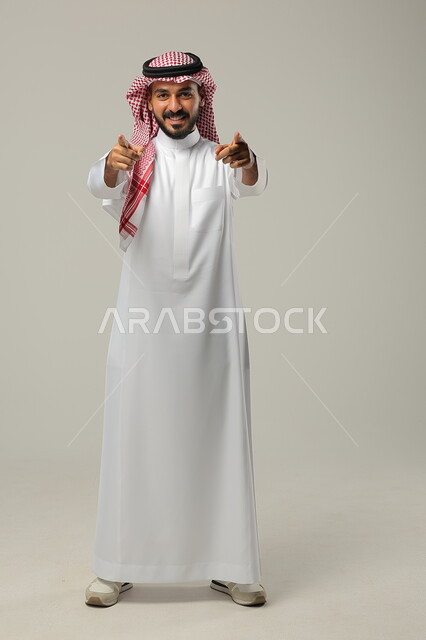 ايماءات التأكيد وتحديد الشيء المقابل, الإشارة الى شخص ما بحركات التواجد هنا, بورتريه لرجل عربي خليجي سعودي يرتدي الثوب التقليدي والشماغ يرفع يديه للأمام بتعابير السرور, صورة الجسم بالطول الكامل, خلفية رمادية