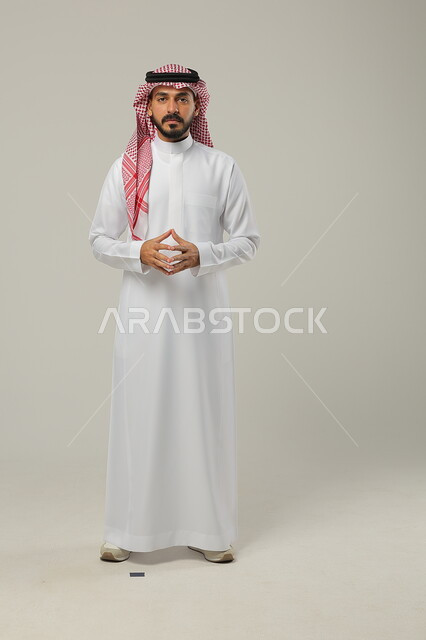 التفكير و التأمل و شرود الذهن والتخطيط للمستقبل, بورتريه لشاب عربي خليجي سعودي يرتدي الثوب التقليدي والشماغ يقوم بملامسة اطراف اصابعه ببعضها، إيجاد حل لمشكلة ما, إيماءات التمعن والتركيز، صورة الجسم بالطول الكامل، خلفية رمادية