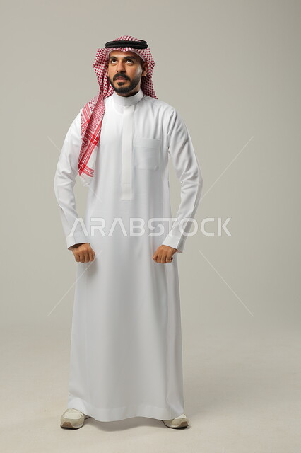الوقوف باستقامة وانضباط، الثقة بالنفس والشعور بالقوة، مفهوم الرجولة و الاهتمام بالمظهر الخارجي، بورتريه لشاب عربي خليجي سعودي يرتدي الشماغ والثوب التقليدي يضع يديه على جانبه و ينظر إلى الاعلى بإيماءات الجدية، صورة الجسم بالطول الكامل، خلفية رمادية
