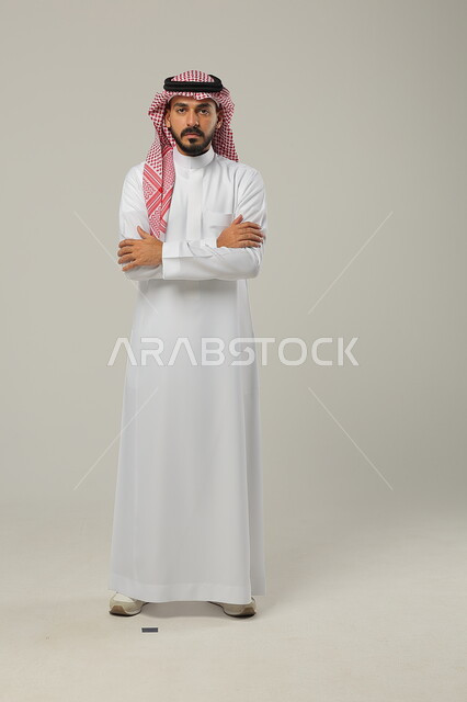 Standing up straight and confident, concept of elegance and masculinity, portrait of a handsome young Saudi Arabian Gulf man wearing a shemagh and traditional dress standing with crossed arms looking at the camera with serious expression, interest in appearance, full body image, gray background
