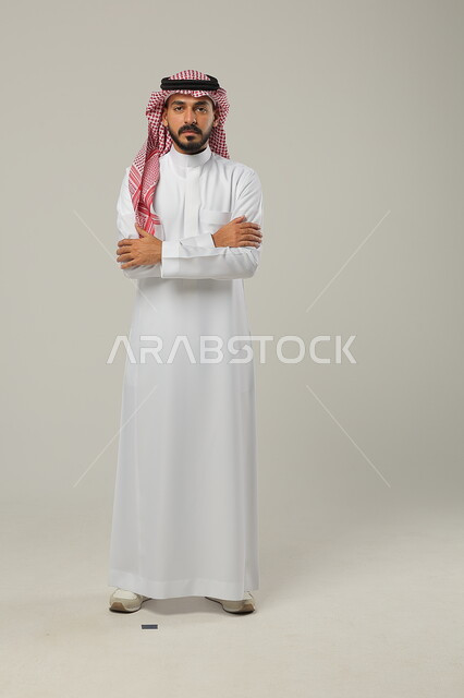  الوقوف باستقامة وثقه بالنفس، مفهوم الاناقه و الرجولة، بورتريه لشاب وسيم عربي خليجي سعودي يرتدي الشماغ و الزي التقليدي يقف بأيدي متقاطعه ينظر إلى الكاميرا بتعابير الجدية، الاهتمام بالمظهر الخارجي، صورة الجسم بالطول الكامل، خلفية رمادية