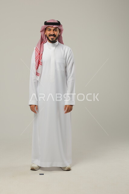 الثقة بالنفس والشعور بالقوة، مفهوم الرجولة والأناقه، الوقوف باستقامة وانضباط، الاهتمام بالمظهر الخارجي، بورتريه لشاب عربي خليجي سعودي مبتسم يرتدي الشماغ والثوب التقليدي يضع يديه على جانبه و ينظر إلى الكاميرا بإيماءات السرور، صورة الجسم بالطول الكامل، خلفية رمادية