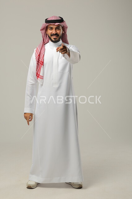 ايماءات التأكيد وتحديد الشيء المقابل, الإشارة الى شيء ما بحركات التواجد هنا, بورتريه لرجل عربي خليجي سعودي يرتدي الثوب التقليدي والشماغ يرفع يديه للأمام بتعابير السرور, صورة الجسم بالطول الكامل, خلفية رمادية